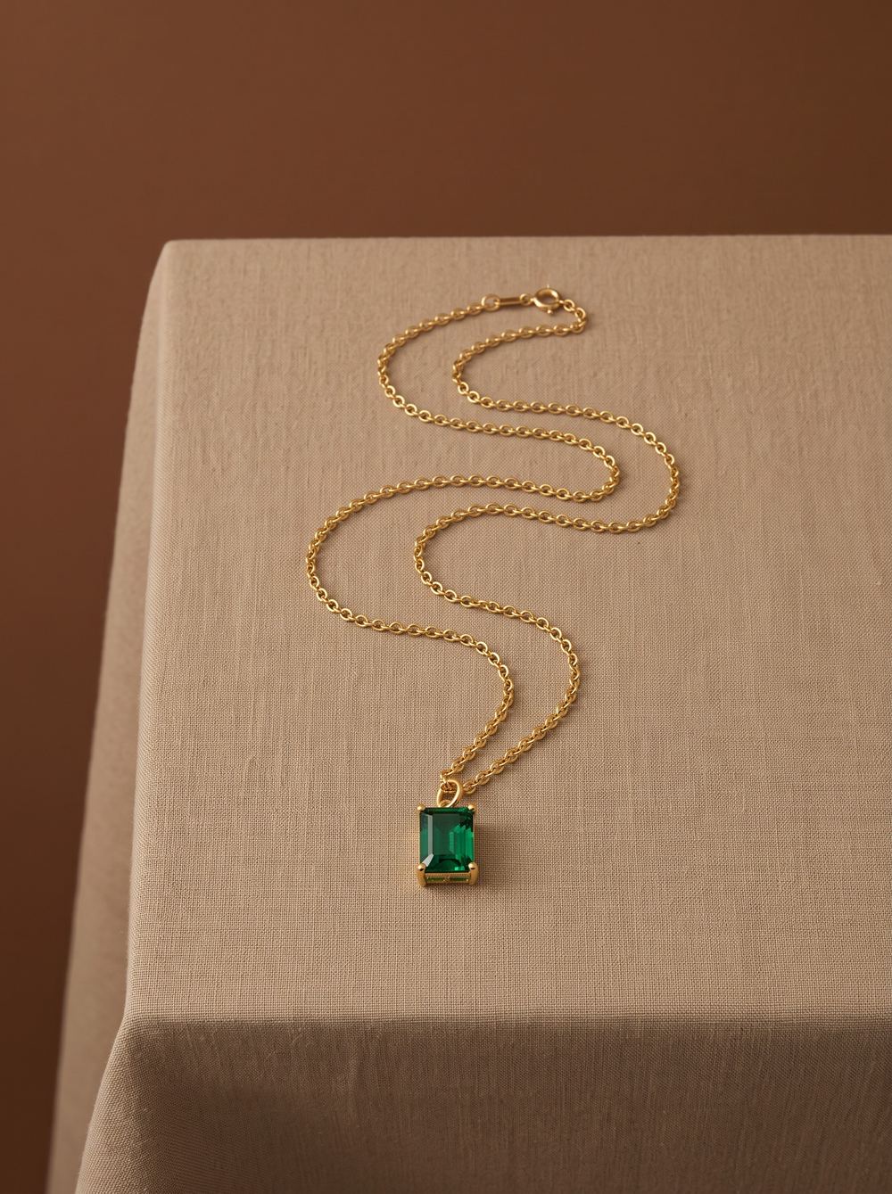 Gold chain with an emerald-cut green pendant laid on linen.